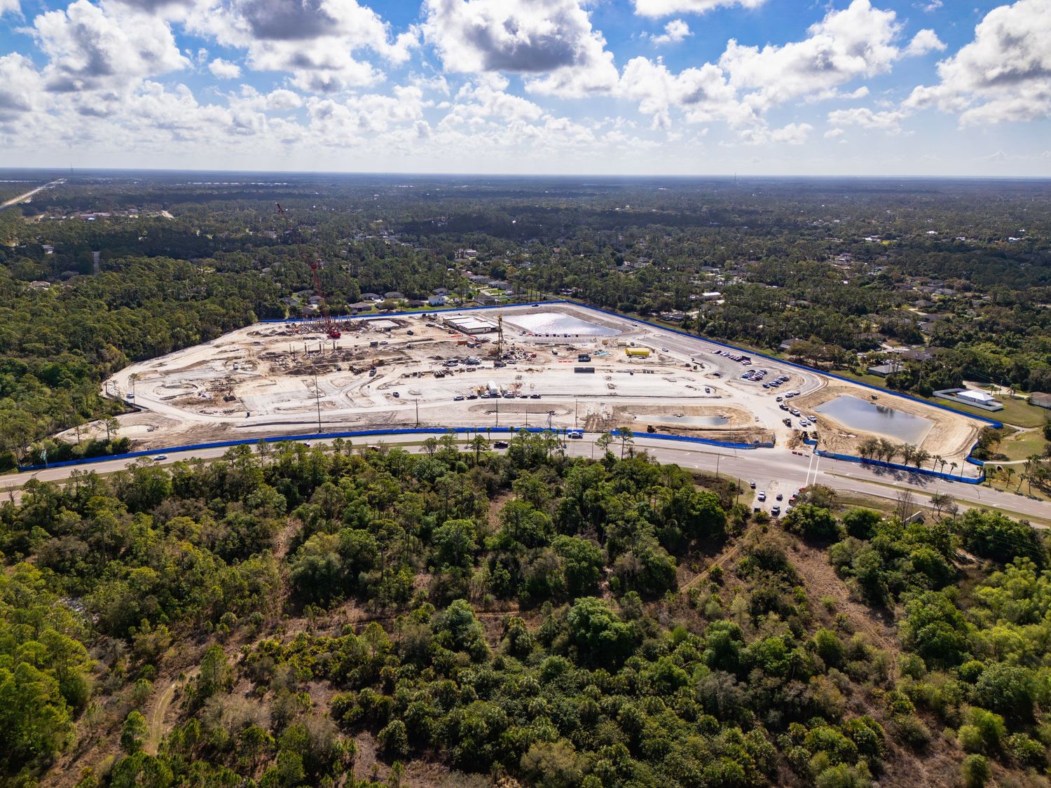 Aerial view of a large construction site surrounded by dense green trees under a partly cloudy sky.
