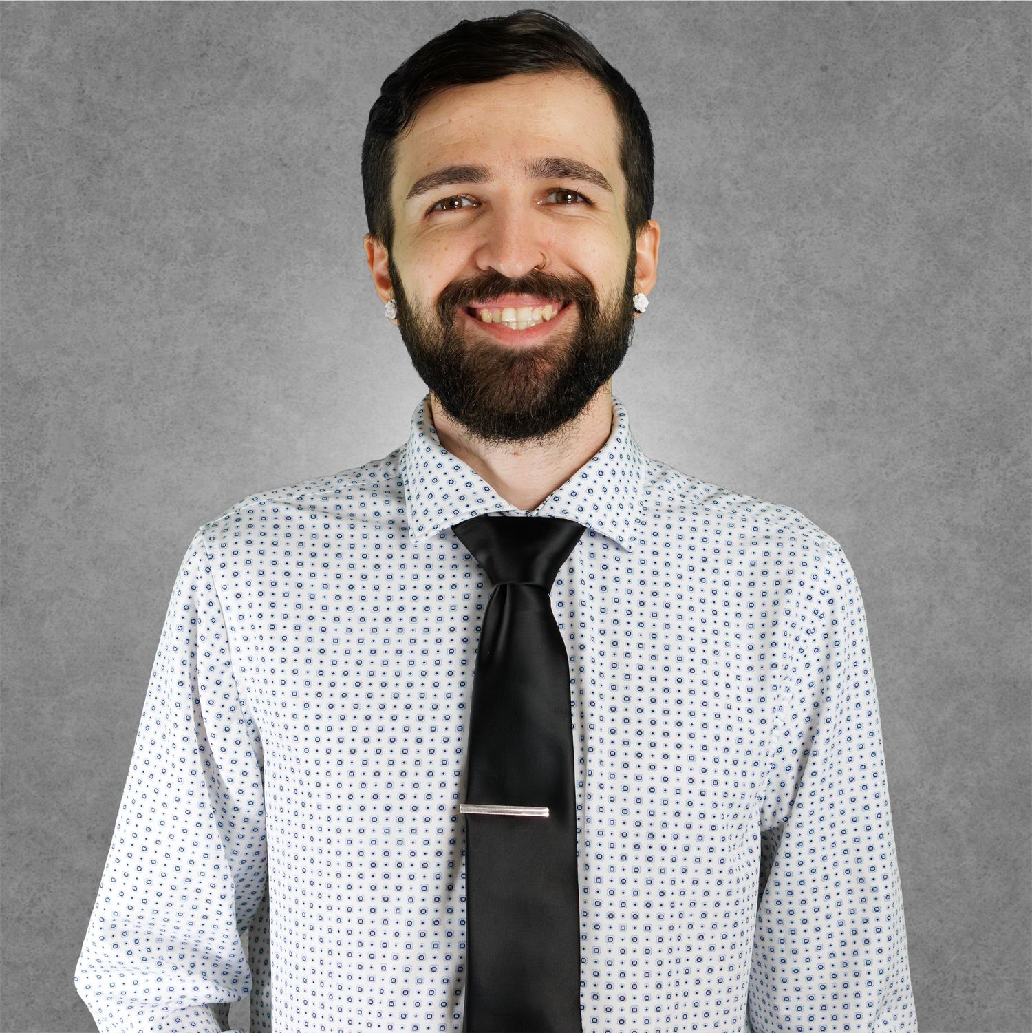 Smiling bearded man wearing a patterned dress shirt and black tie with tie clip against gray background.