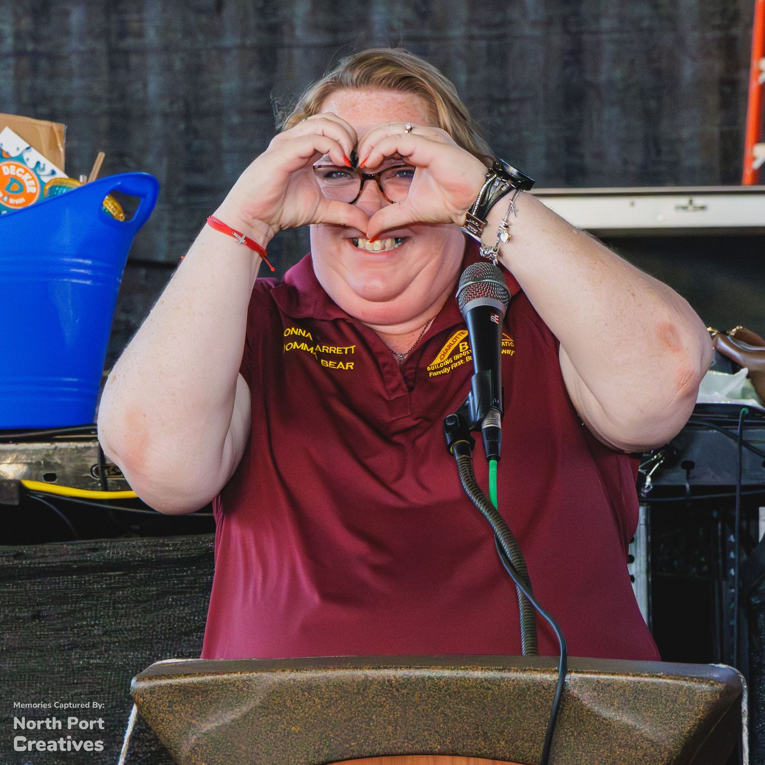 Woman in maroon polo making heart shape with hands at podium during outdoor event.