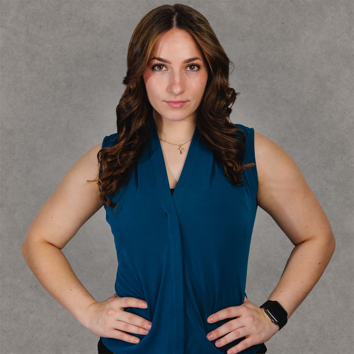 Professional woman in teal sleeveless top with hands on hips, posing confidently against gray background.