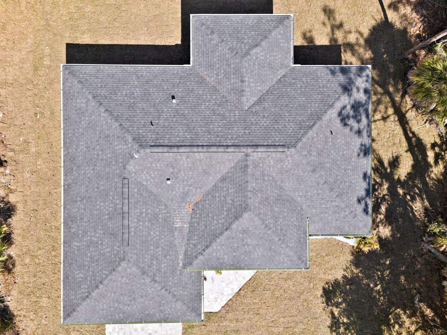 Aerial view of a gray asphalt shingle hip roof on a residential home surrounded by dry grass and trees.