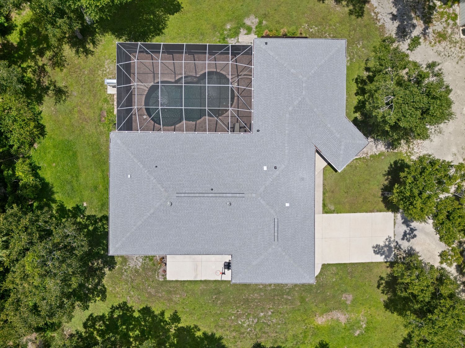 Aerial view of a gray hip roof home with screened pool enclosure surrounded by lush green trees and landscaping.