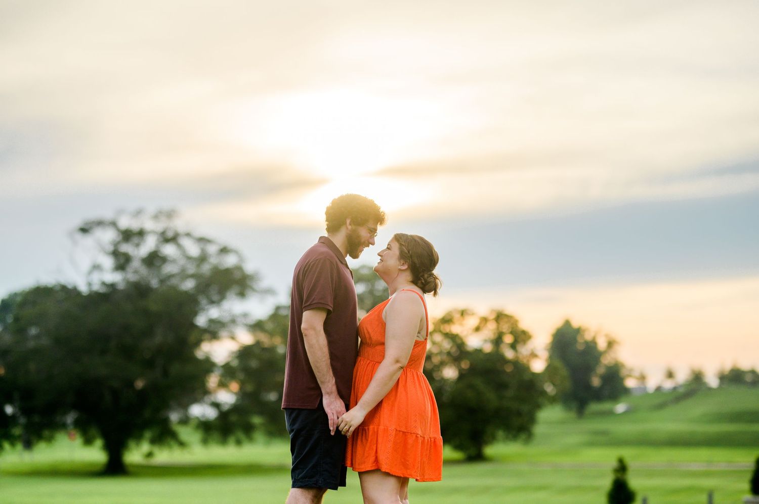 A romantic sunset moment captured between a couple in an orange dress and dark shirt on a grassy hill. Knoxville Couple Photographer