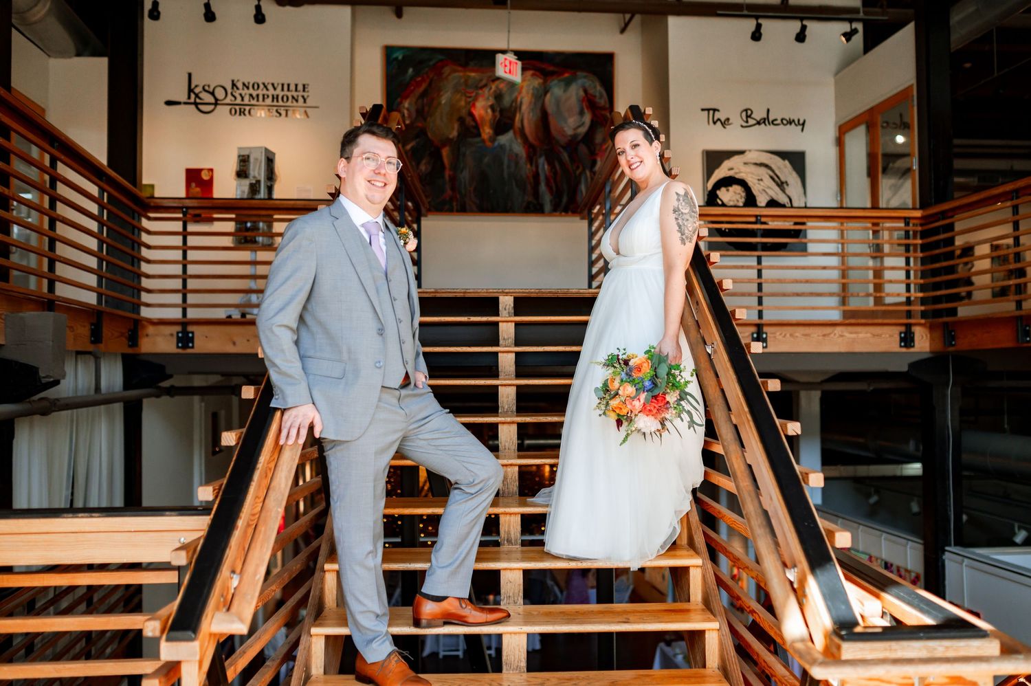 A couple in wedding attire poses joyfully on wooden stairs inside a modern industrial-style venue.