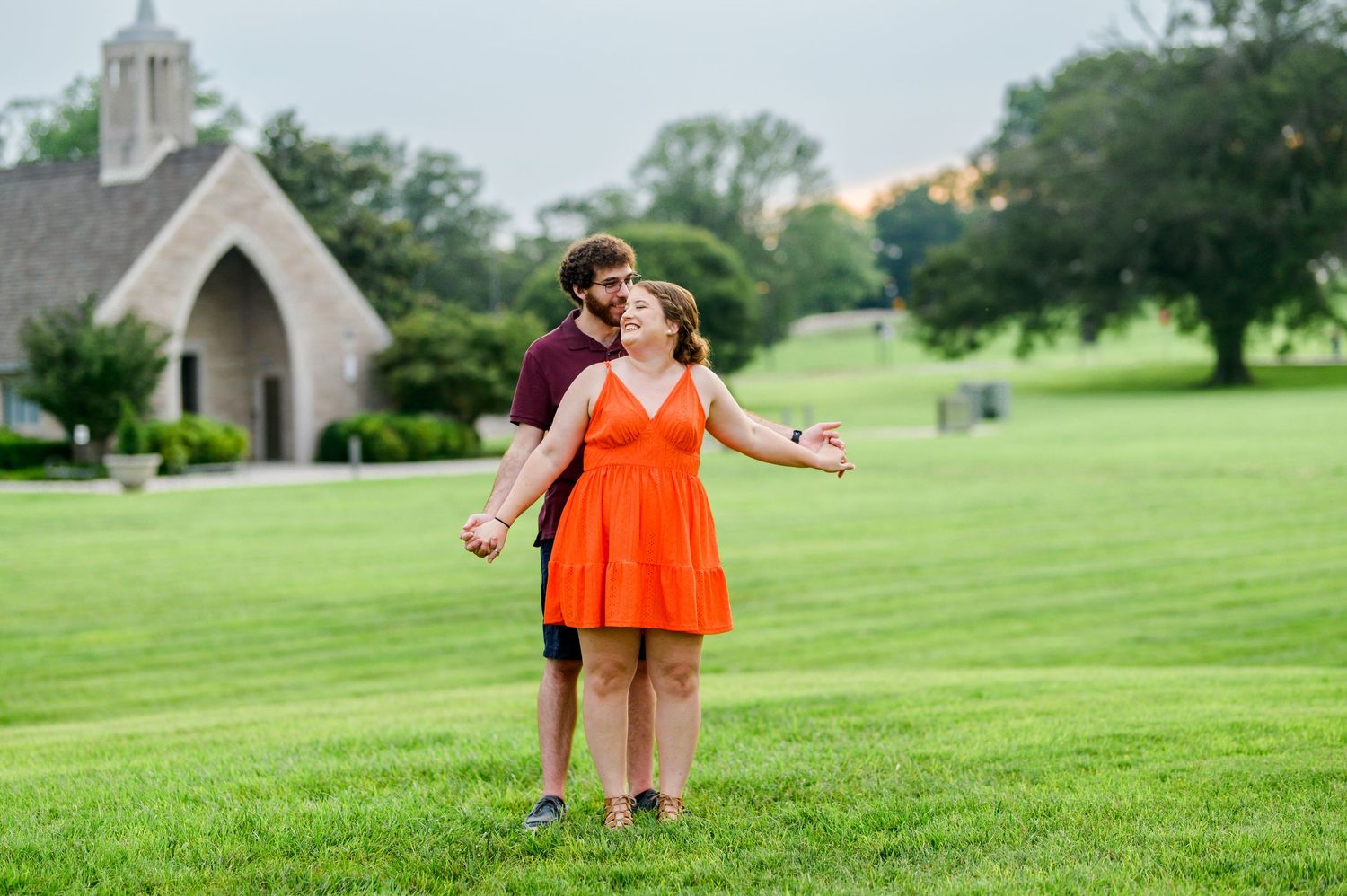 A couple joyfully poses on a grassy lawn with a church building visible in the background during golden hour. Knoxville Couple Photographer