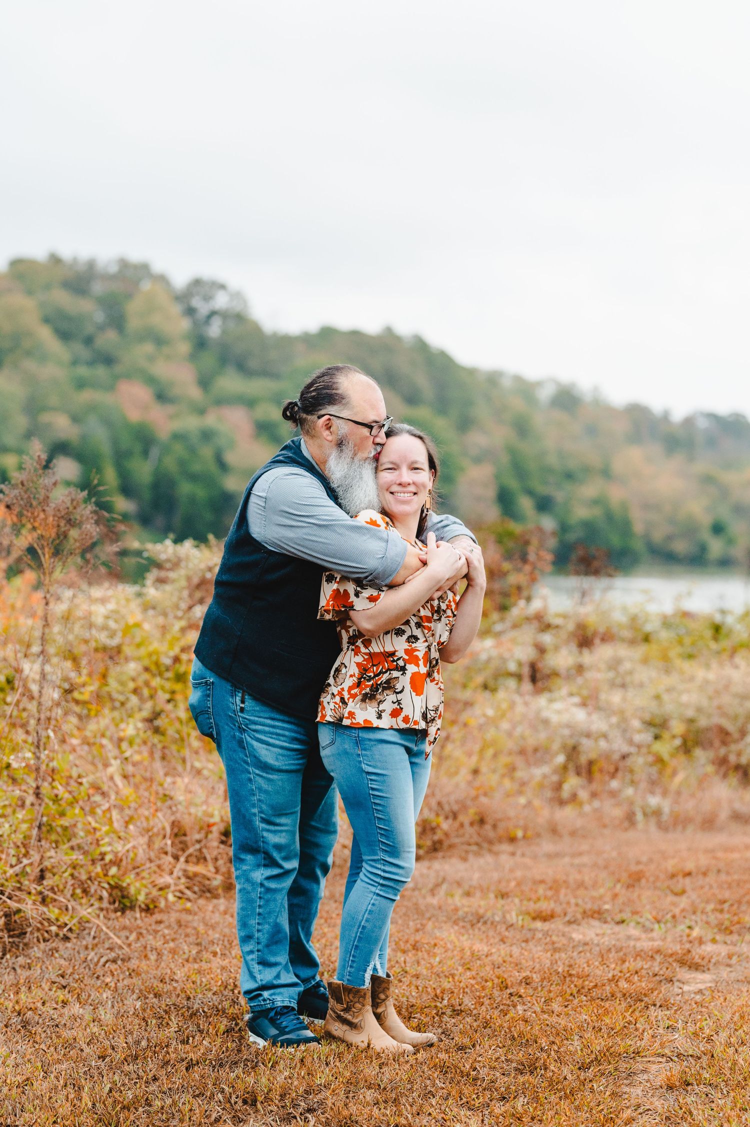 A couple shares a joyful embrace while standing in an autumn field with fall foliage in the background. Knoxville Couple Photographer