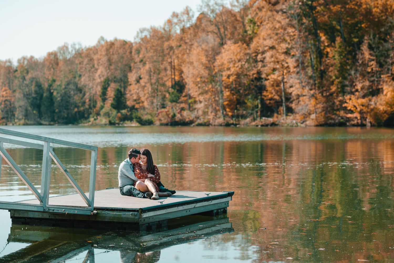 A couple sits close together on a dock over calm waters surrounded by autumn trees. Knoxville Couple Photographer. Melton Hills Park