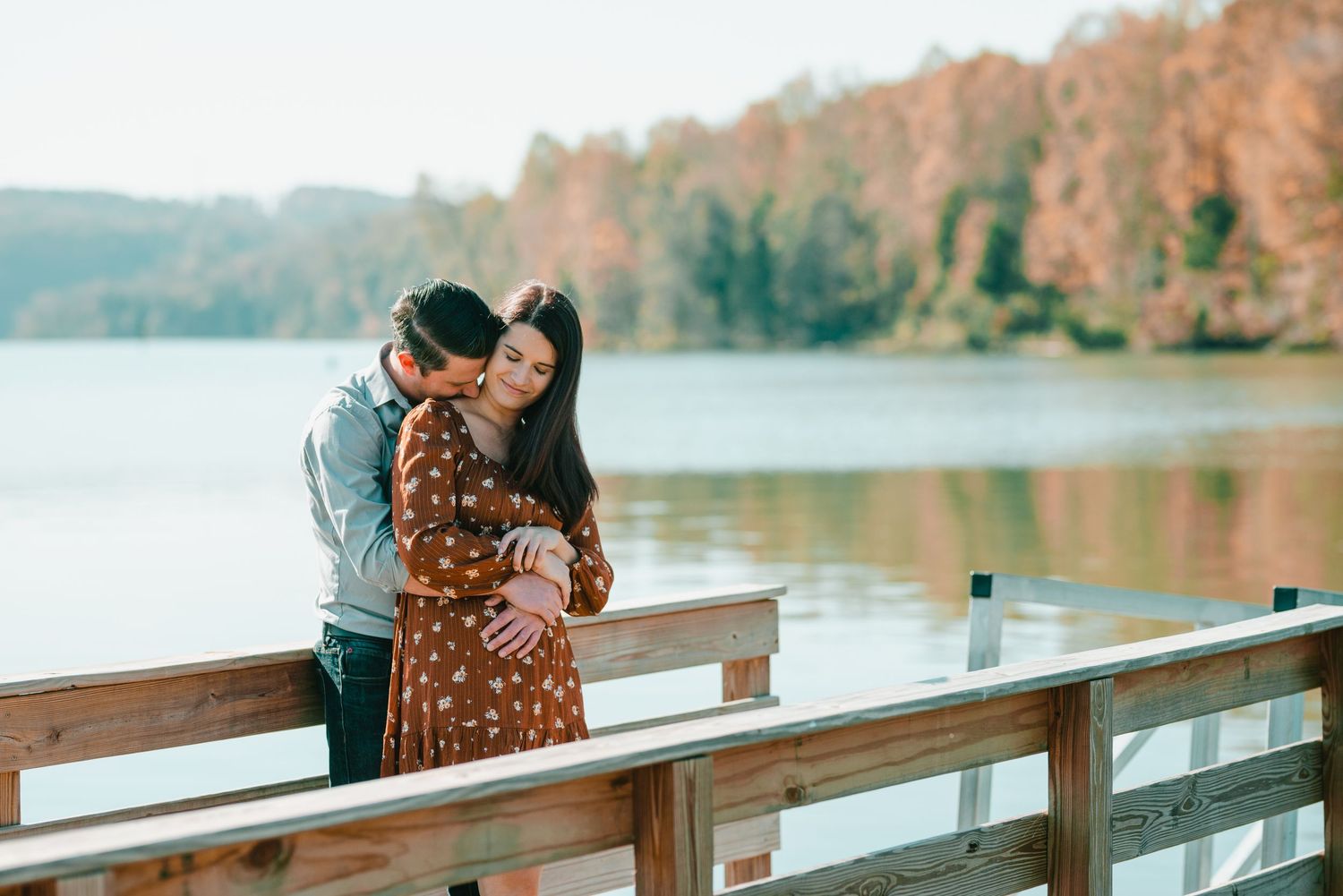 A couple shares a romantic moment on a wooden dock overlooking a lake with fall colors. Knoxville Couple Photographer