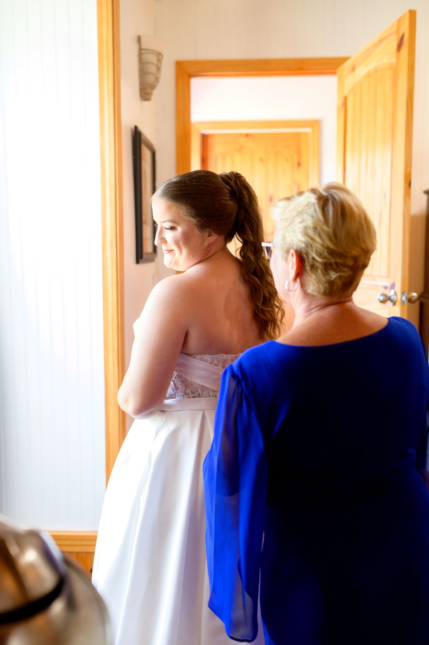 A bride in white and woman in blue prepare for the wedding day in a sunlit room. Knoxville Wedding Photographer