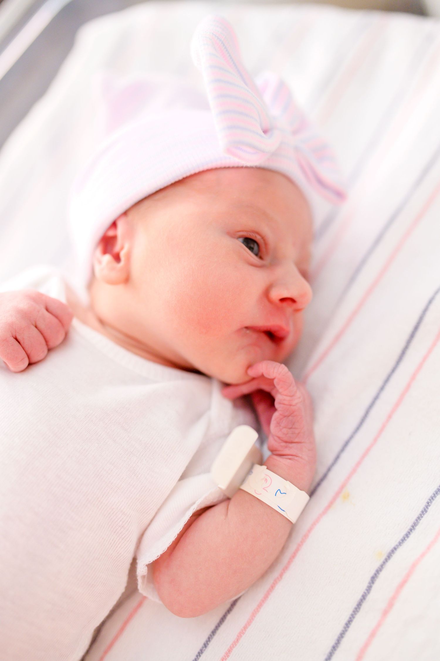 A newborn baby wearing white clothing lies on striped bedding while sucking their thumb. Knoxville Newborn Photographer UT Medical Center
