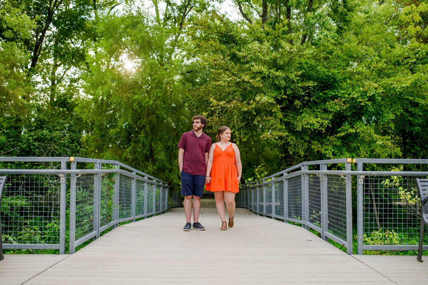 A couple walks hand in hand across a metal bridge surrounded by lush green trees. Knoxville Couple Photographer. Lakeshore Park