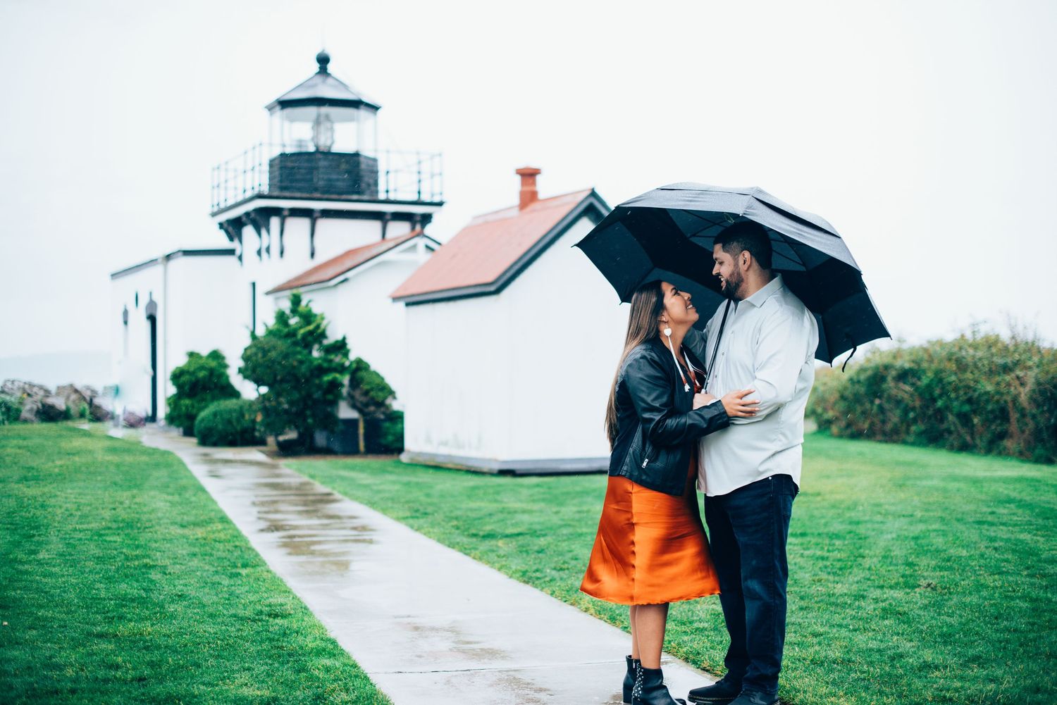 Couple sharing romantic moments under umbrellas near a lighthouse on a rainy day. Knoxville Couple Photographer