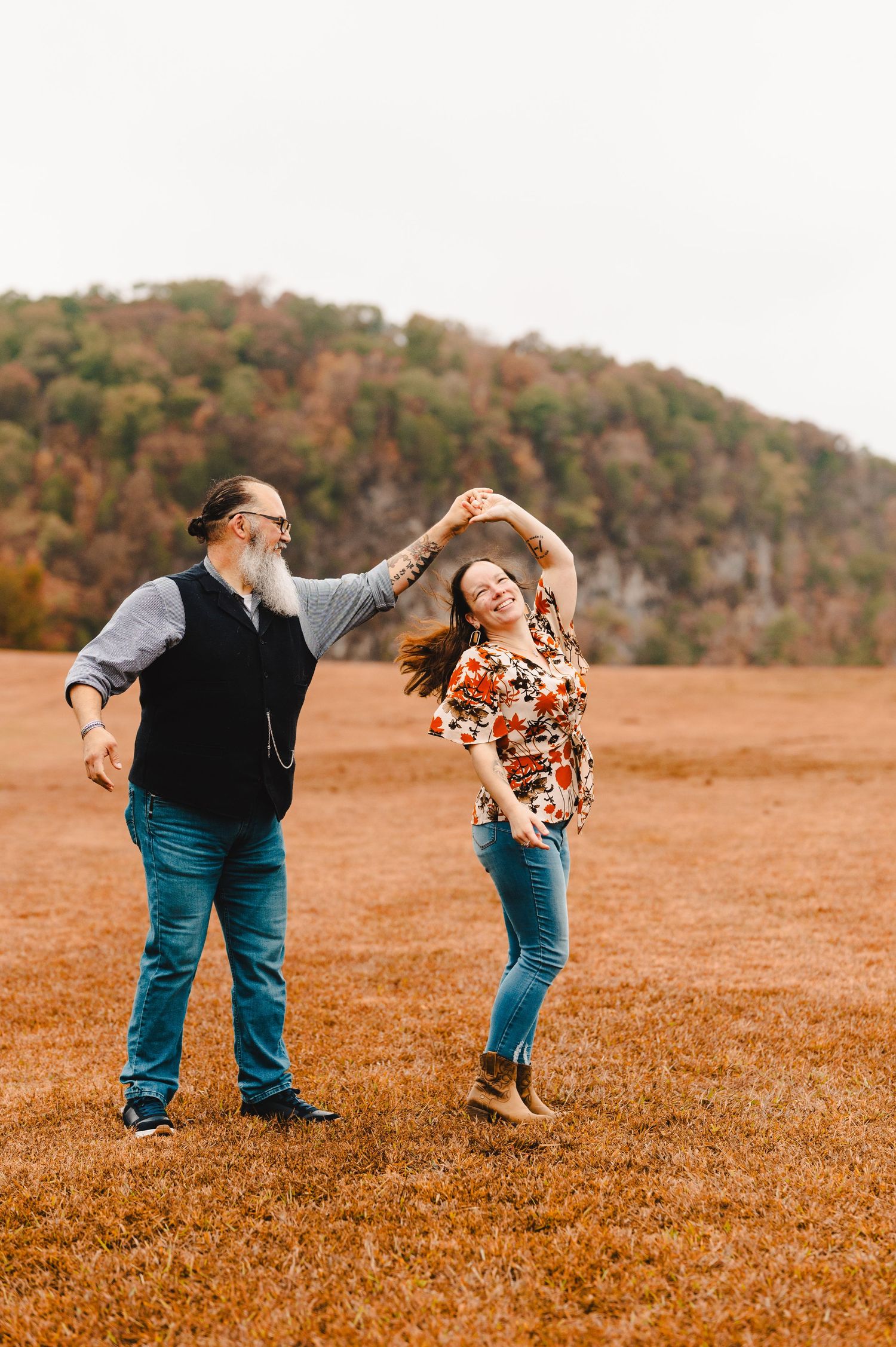 A couple dances playfully in a field against a backdrop of autumn trees on a hillside. Knoxville Couple Photographer