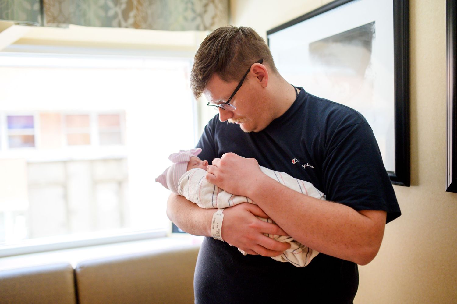 A tender moment as a parent in a navy shirt cradles a newborn baby wrapped in a blanket near a window. Knoxville Newborn Photographer UT Med