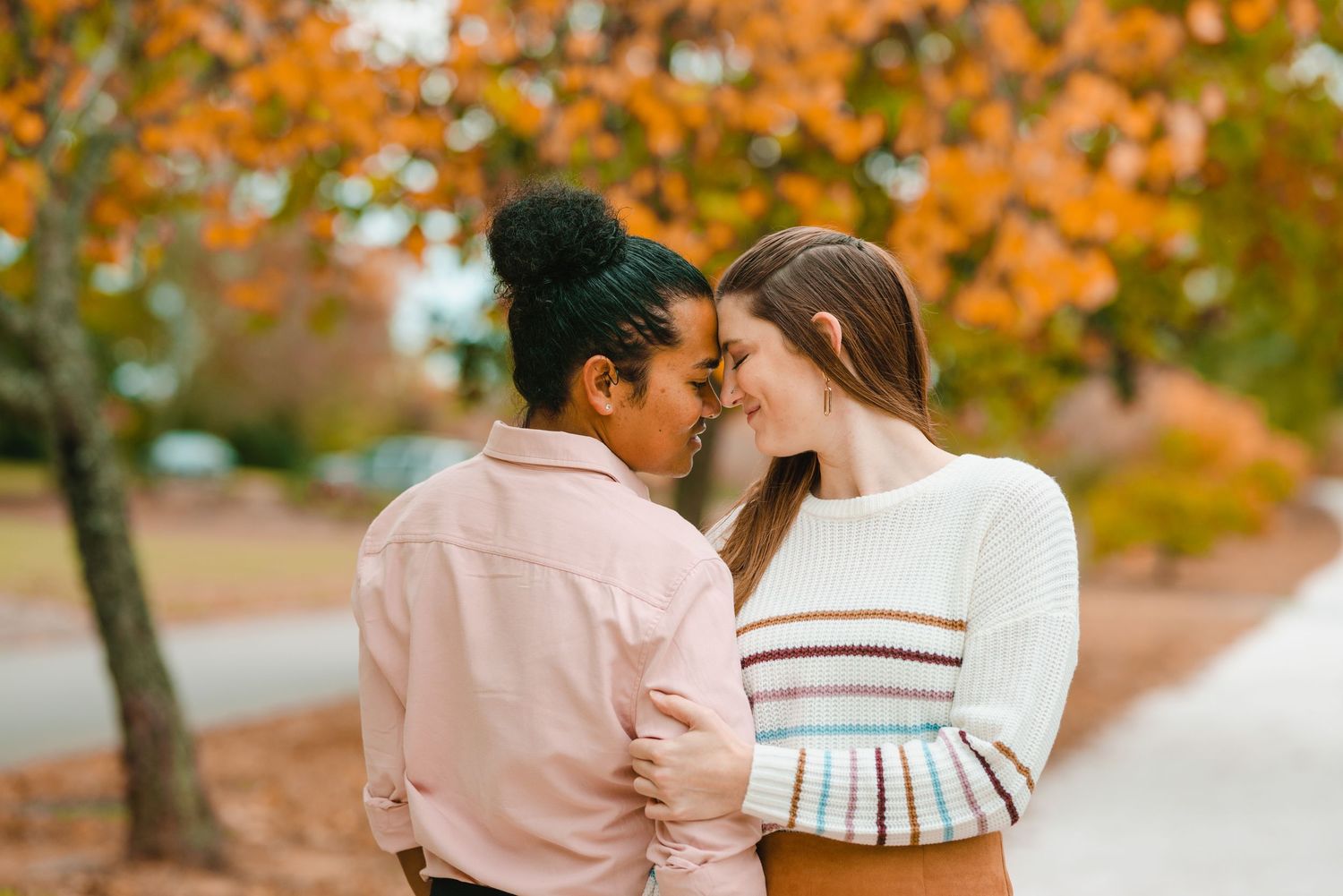 Two people share a romantic moment surrounded by vibrant orange fall foliage in a park setting.