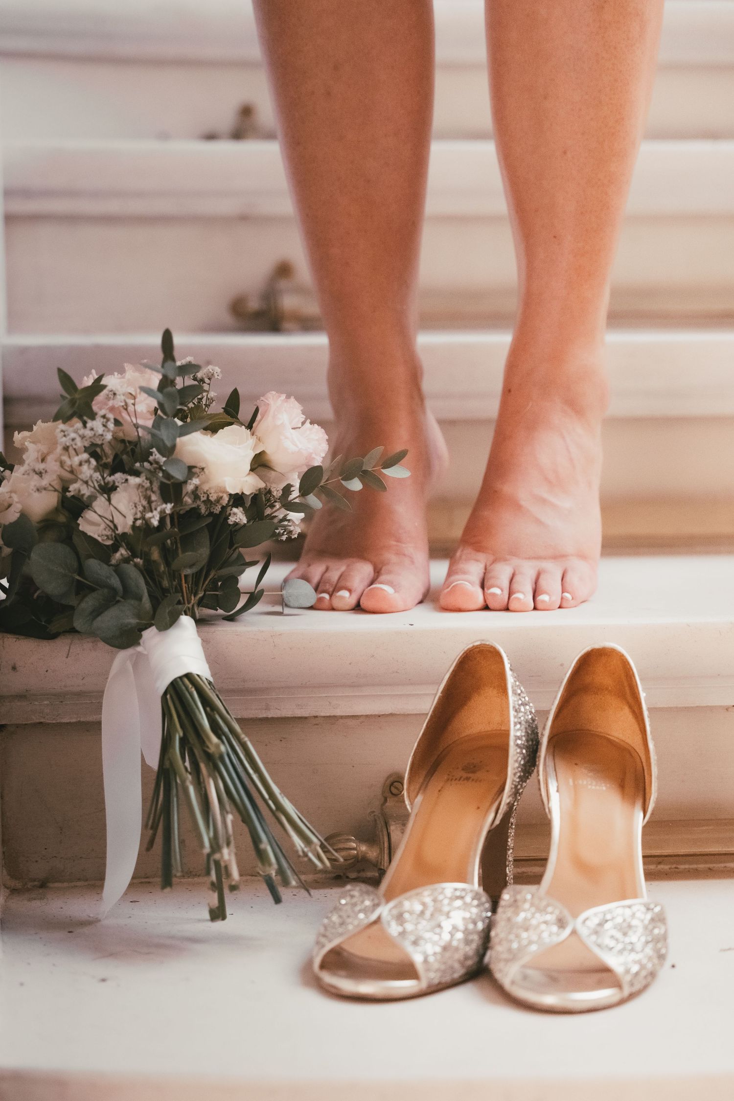 Barefoot bride standing on white stairs with elegant crystal heels and white floral bouquet tied with ribbon.