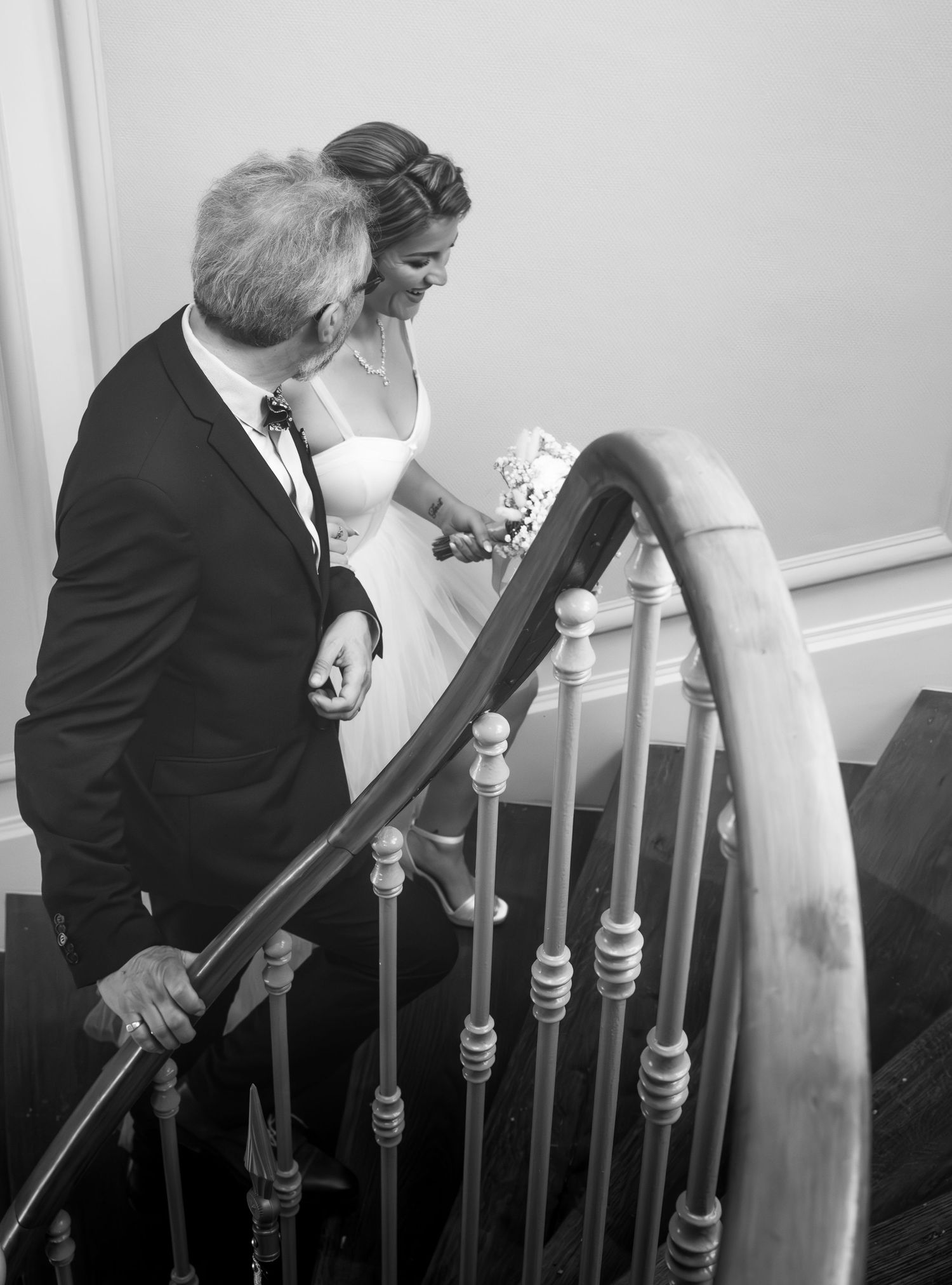La futur mariée au bras de son beau père dans une tenu formelle monte ensemble un élégant escalier incurvé en photographie en noir et blanc