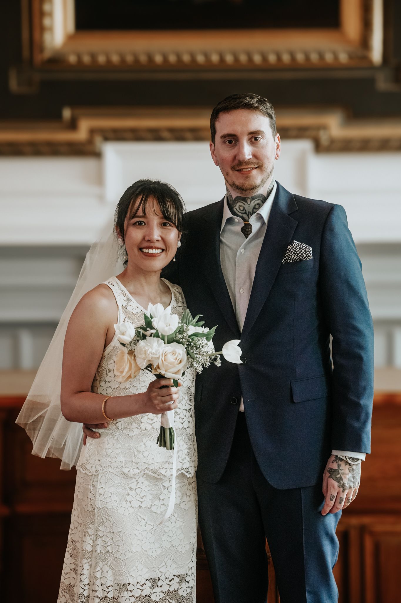 A bride and groom pose together in a traditional church setting with elegant wedding attire and white floral bouquet.