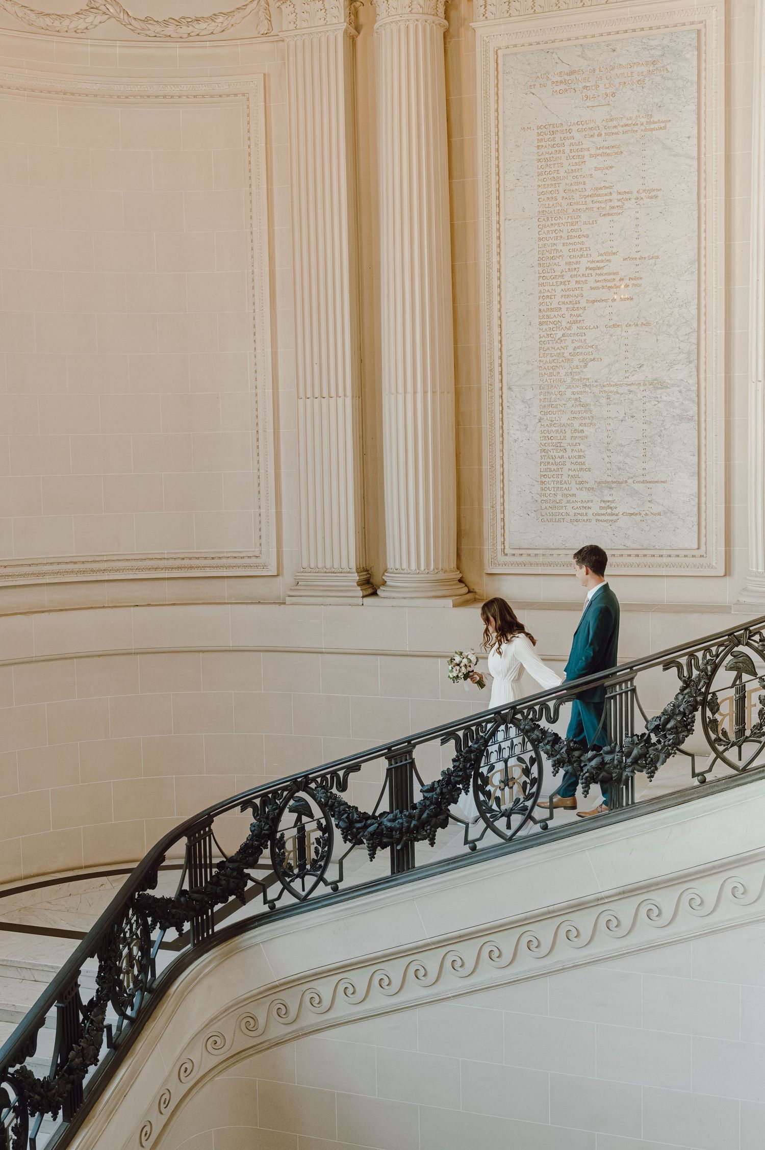 A bride and groom walk down an ornate staircase with iron railings inside an elegant neoclassical building.