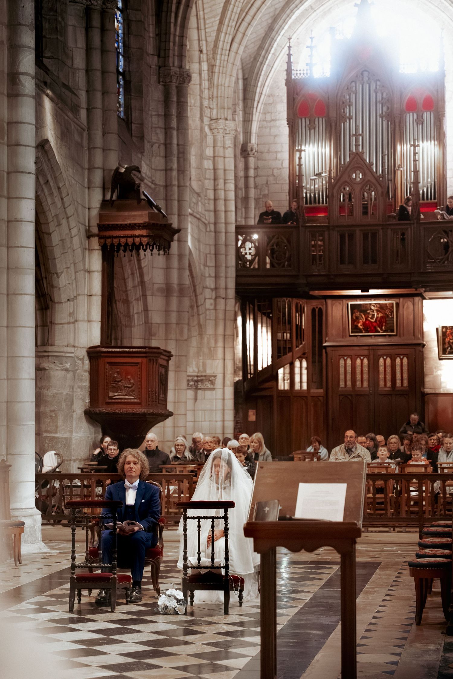 Bride and groom seated in a grand cathedral during a wedding ceremony with ornate wooden organ pipes in background.