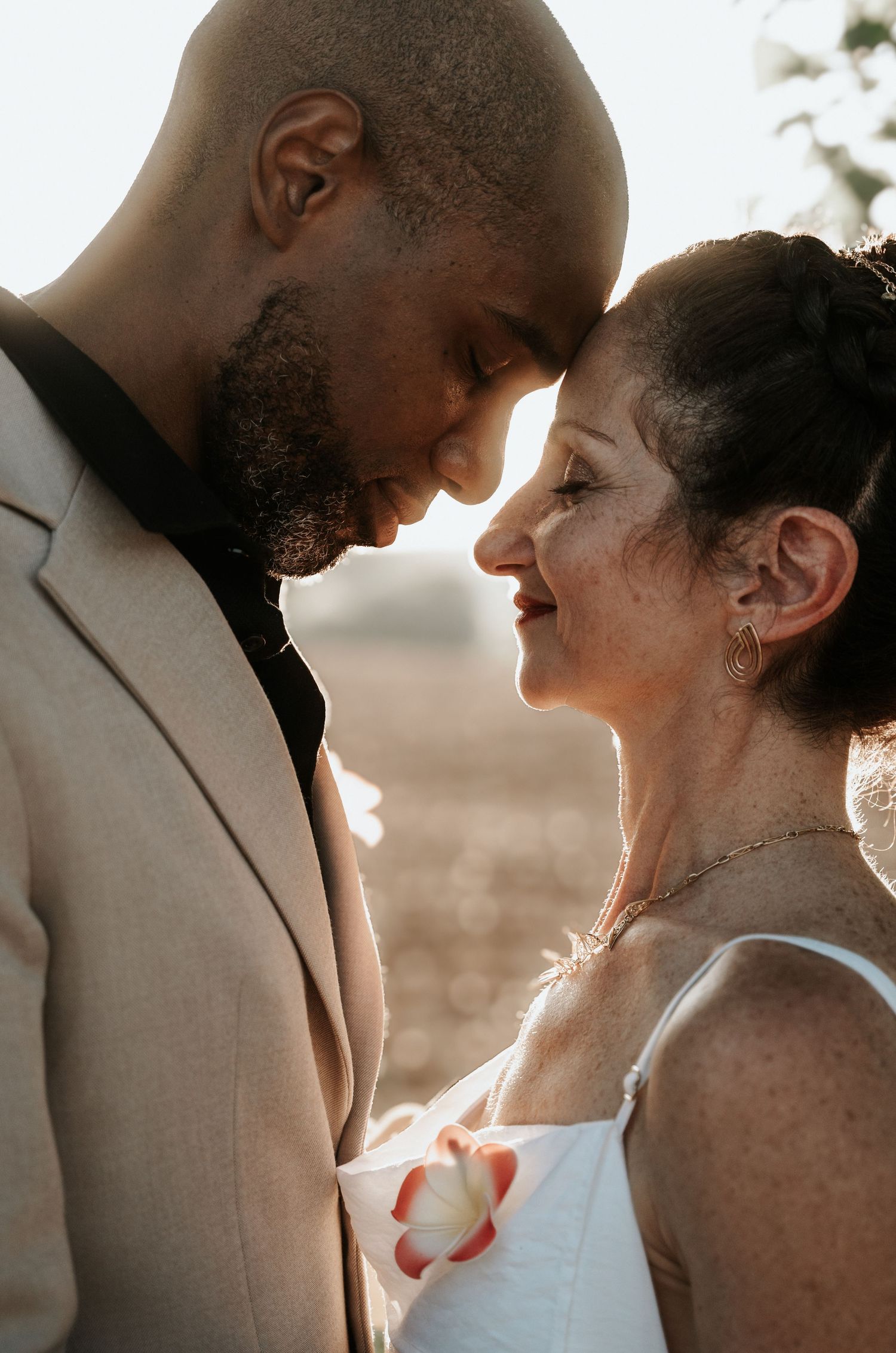 A romantic couple shares an intimate moment in sunlit wedding attire, with the groom in beige and bride in white.