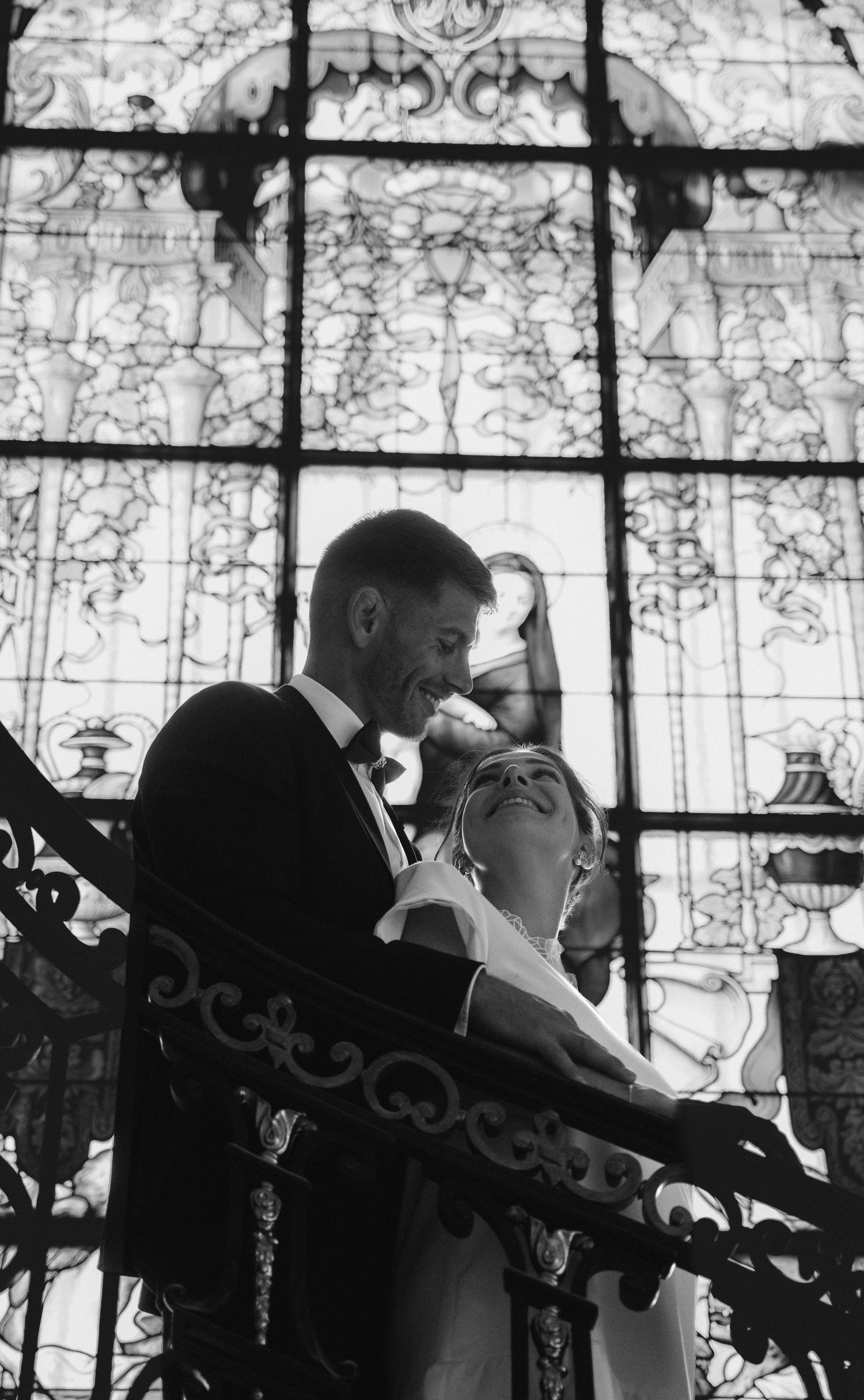 Black and white wedding photo of smiling couple embracing on ornate staircase before stunning stained glass window.