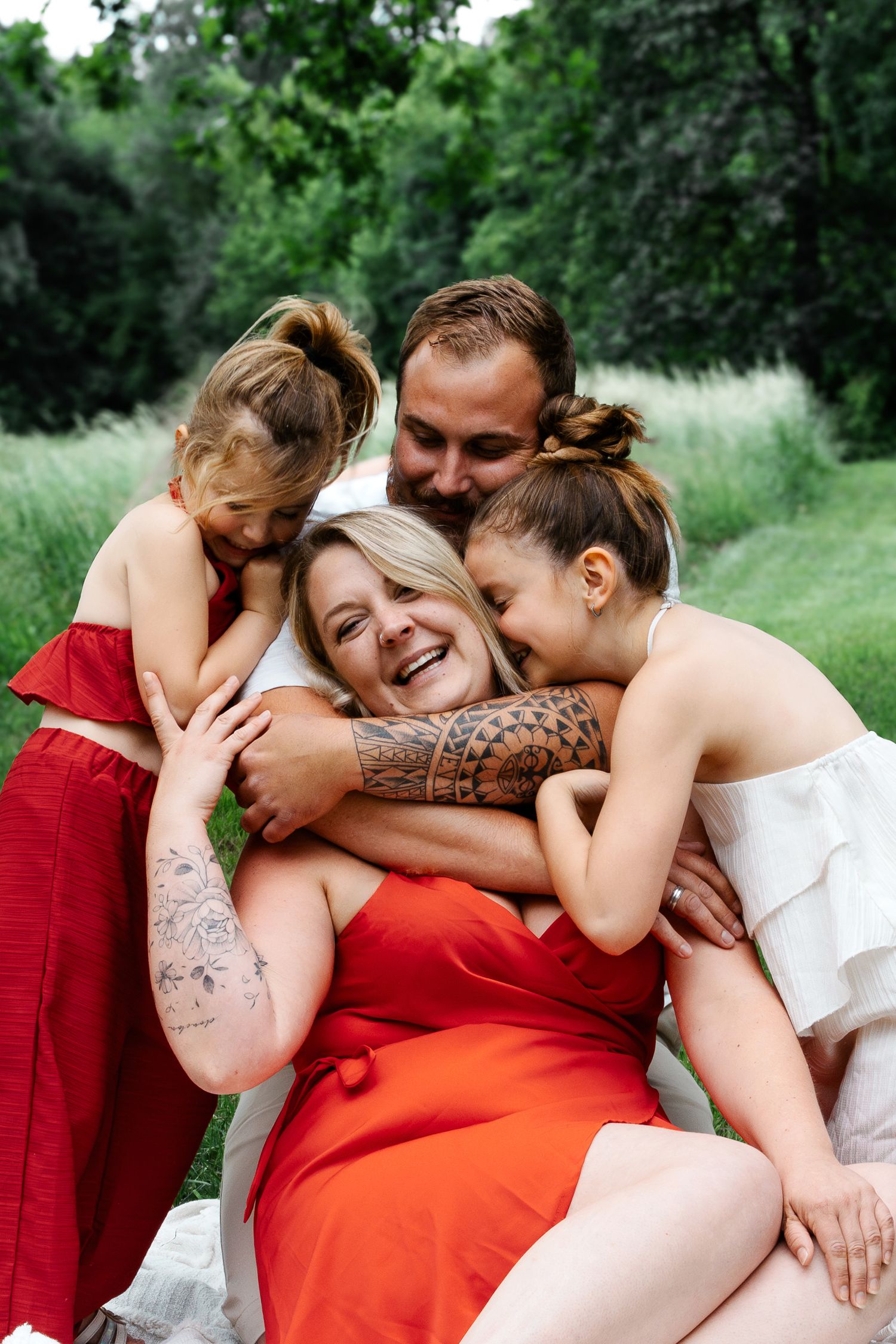 Happy family hugging outdoors, parents and two girls in red and white dresses sharing a joyful embrace in a green park.