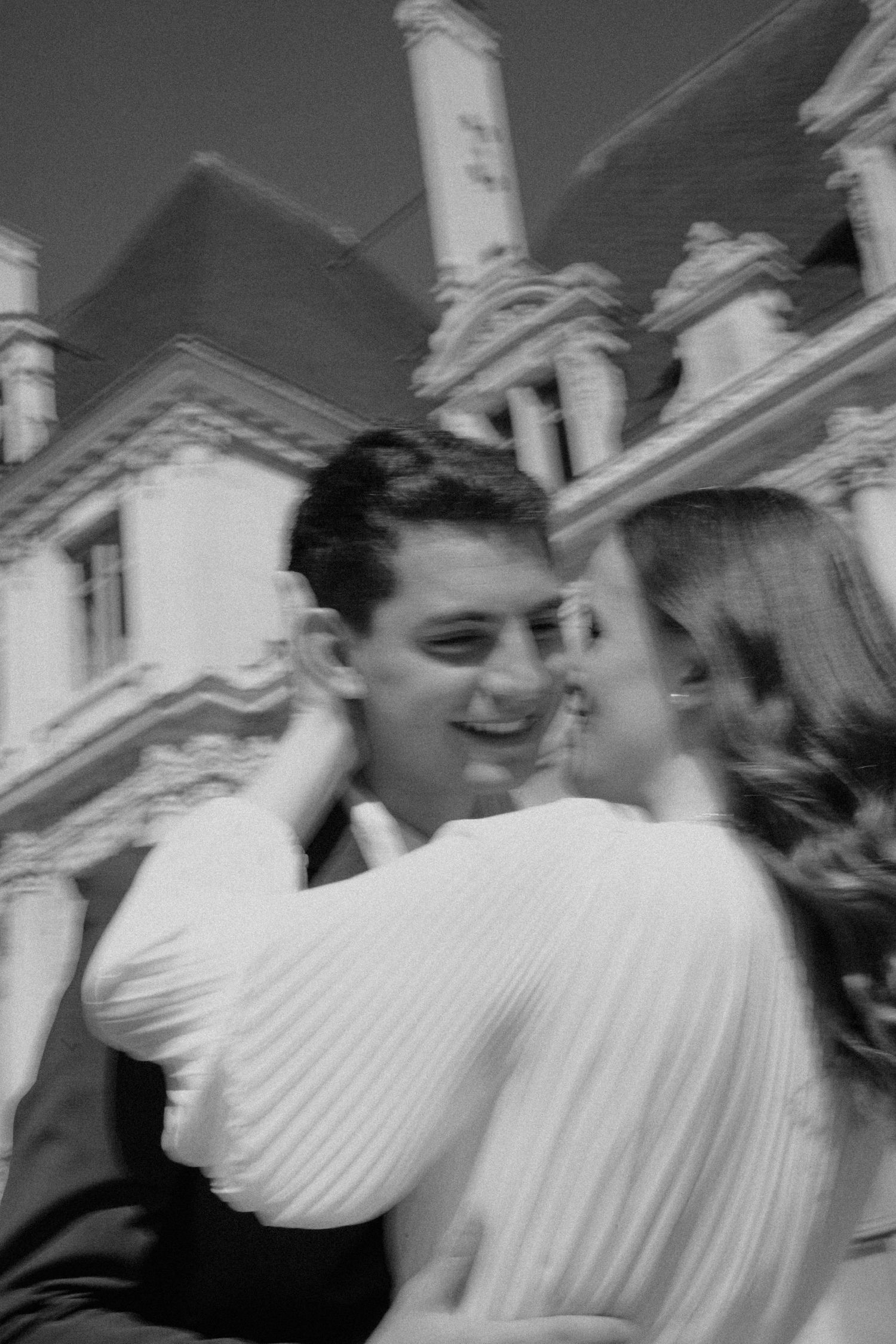 Black and white photo of a smiling couple embracing outside a grand historic building.