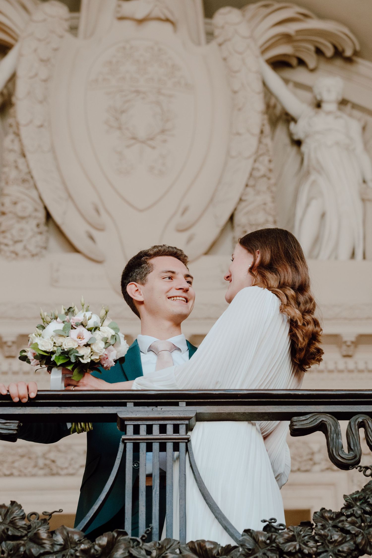 Happy groom holding bouquet with bride on elegant balcony with classical sculptures backdrop.