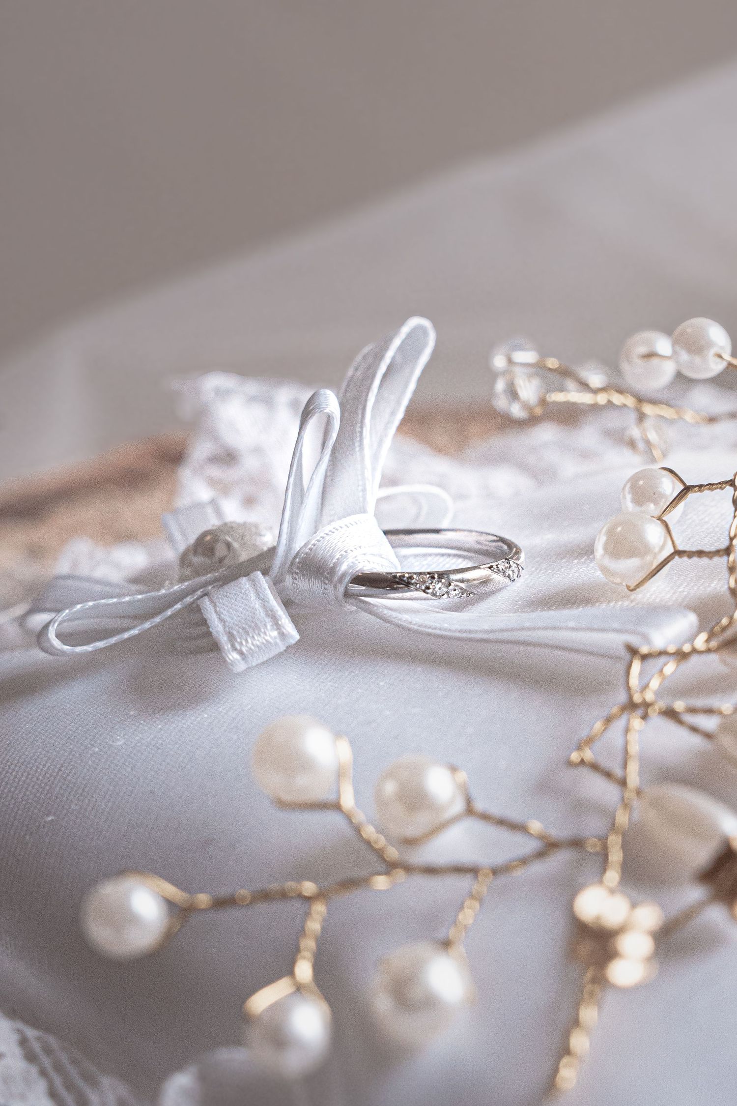 Wedding rings tied with white ribbon rest on silk fabric alongside delicate pearl and gold branch decorations.