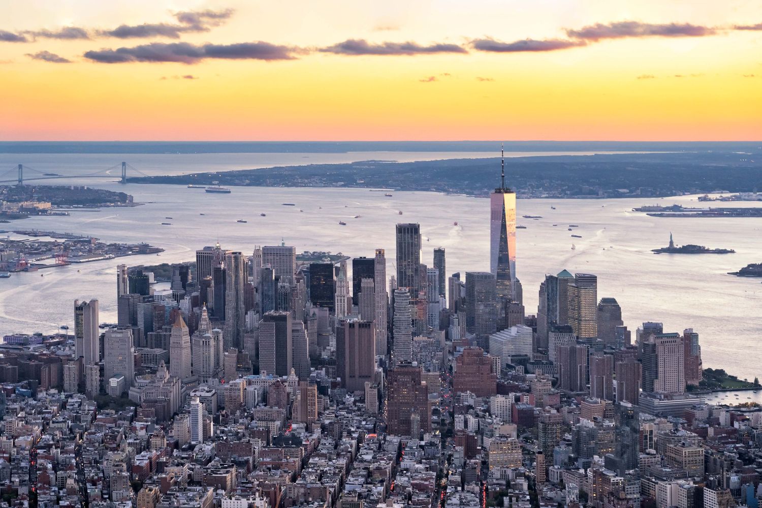 Panoramic sunset view of downtown Manhattan's skyline with harbor and orange sky from a helicopter.