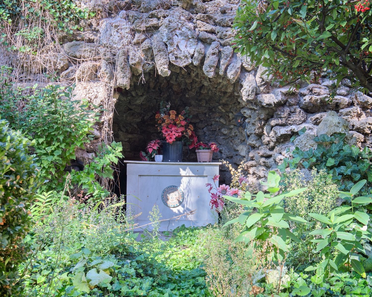 Stone grotto shrine with red flowers and a decorative plaque, surrounded by lush green garden foliage.