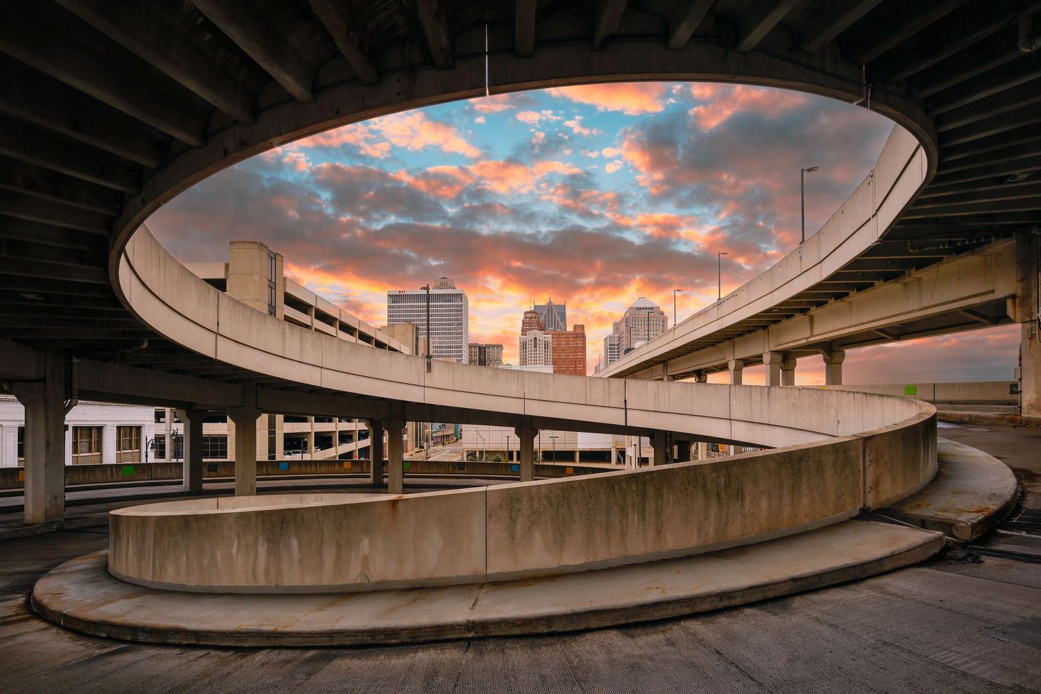 Curved Cobo concrete parking lot ramp under a dramatic sunrise sky with Detroit city buildings in the distance.