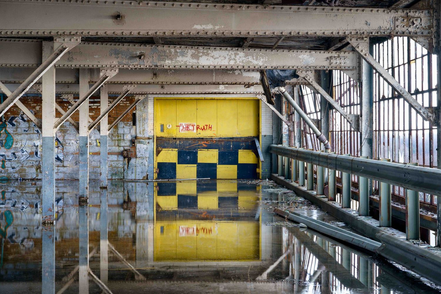 Abandoned Cadillac Stamping plant in Detroit with a yellow elevator door surrounded by metal beams with reflection from water on the floor.