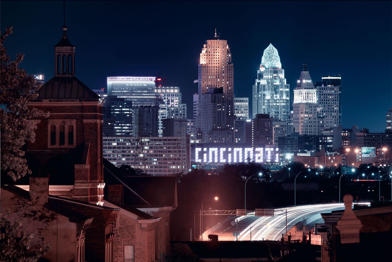 Cincinnati skyline at night with illuminated buildings and highway lights creating light trails.