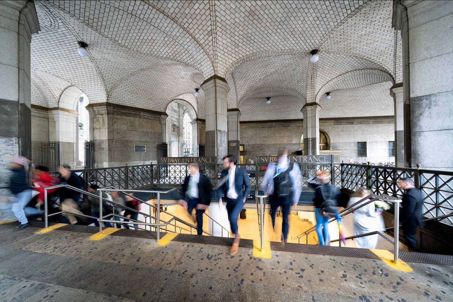 Manhattan subway stairs with geometric ceiling patterns and blurred people coming up the stairs exiting the station.