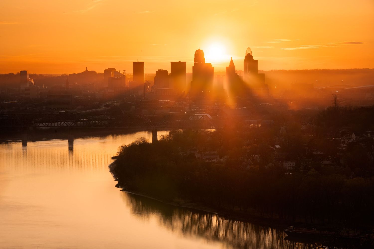 Dramatic orange sunrise over Cincinnati's skyline with sun rays breaking through foggy clouds with the Ohio river in the foreground.