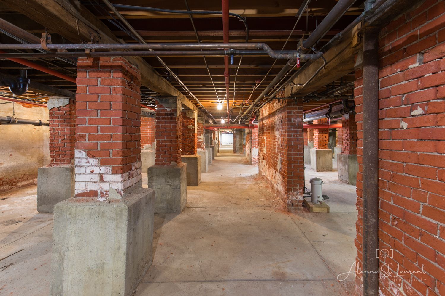 Brick columns line a spacious basement corridor with exposed pipes and concrete floors in an industrial building.