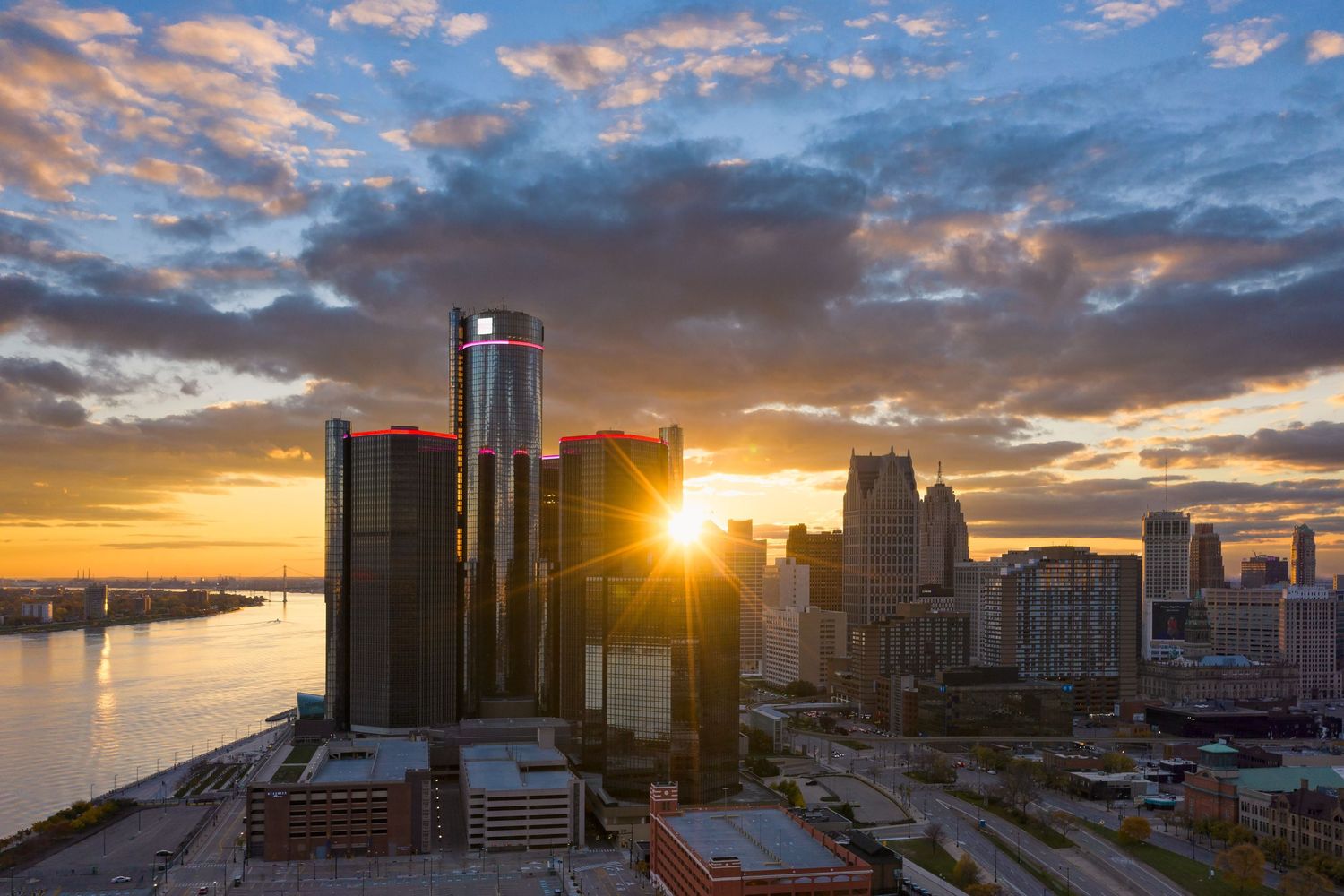 Detroit city skyline at sunset with dramatic clouds and golden light starburst between buildings of the Renaissance Center.