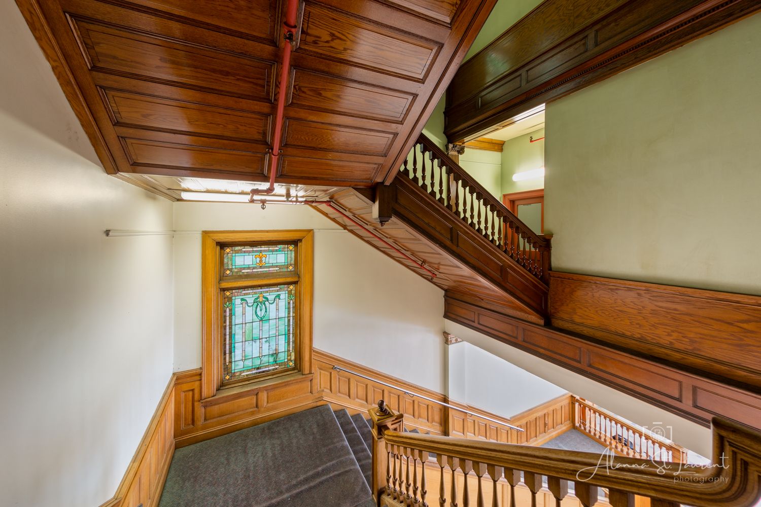 Elegant Victorian staircase with rich wood paneling, ornate balusters, coffered ceiling, and stained glass entry door.