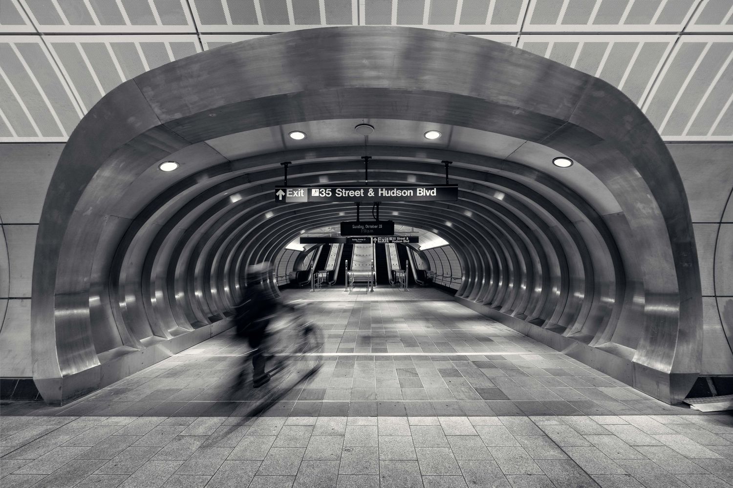 NYC subway station walkway with arched ceiling and dramatic lighting creating symmetrical patterns with man on bike caught in movement.