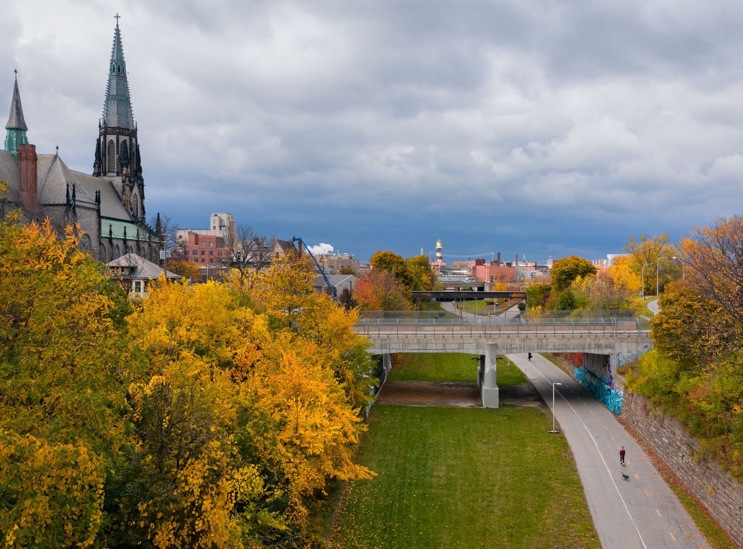 Aerial view of the Dequindre Cut greenway with fall foliage and St Joseph church against an overcast sky with a person walking on the path.