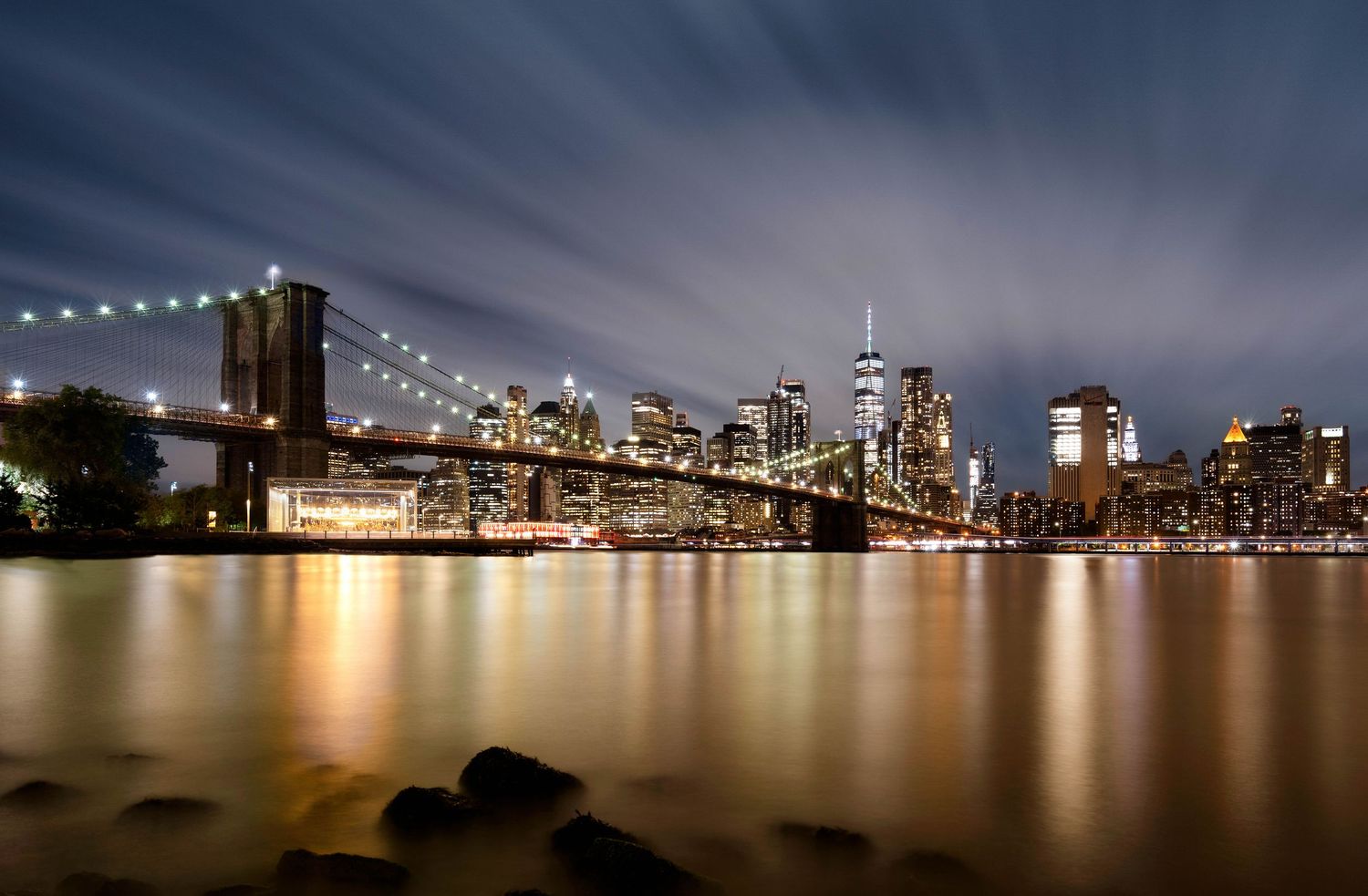 Night panorama of Manhattan city skyline with Brooklyn bridge and twinkling lights reflecting on dark waters with long exposure clouds.
