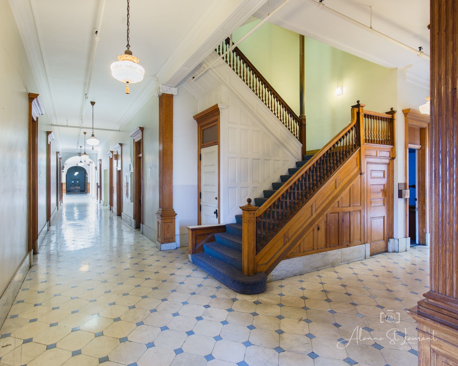 Historic building hallway with ornate wooden staircase, decorative tile floors, tall ceilings, and arched corridor with pendant lighting.