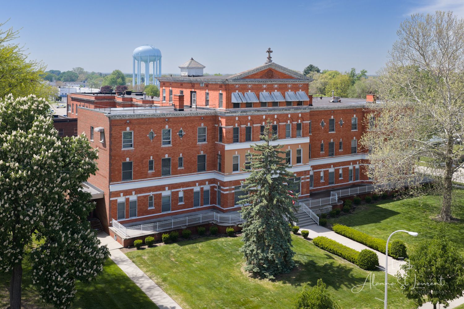 Aerial view of a historic red brick building with manicured lawns and a water tower in the background on a sunny day.