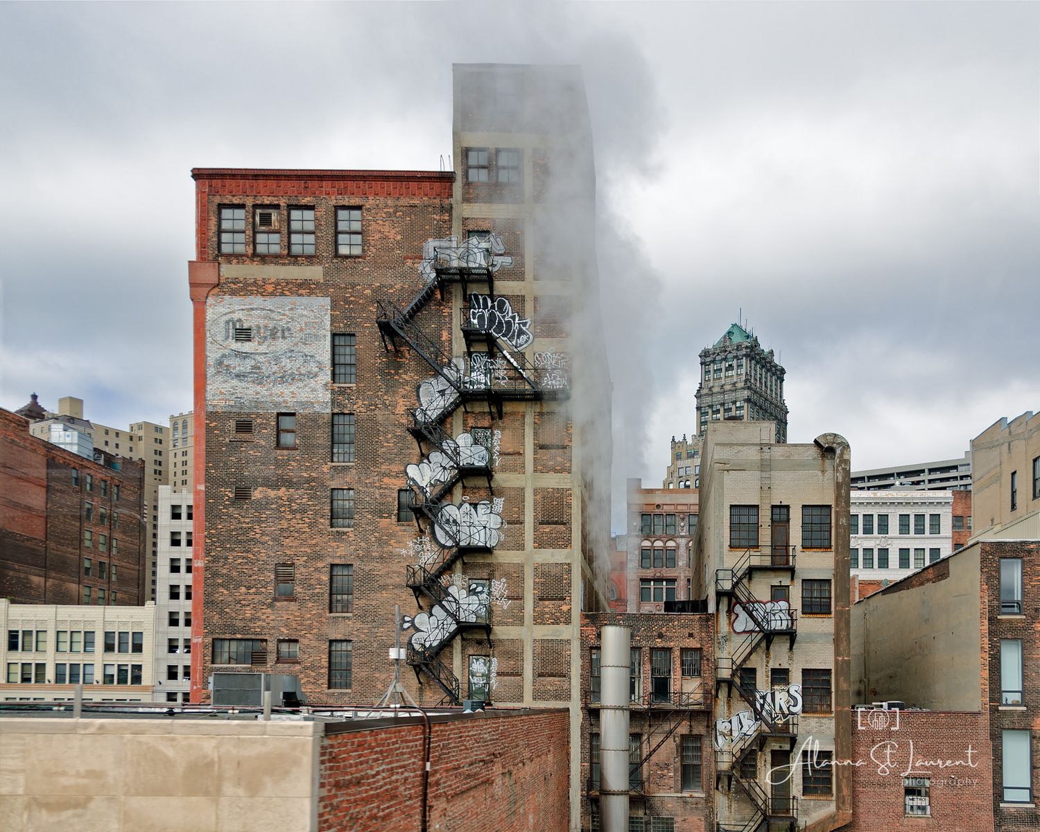 Historic brick buildings with fire escapes and steam rising between them under an overcast sky in an urban cityscape.