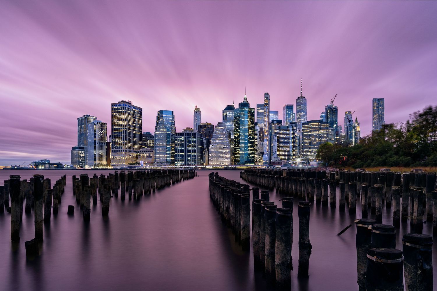 Purple sunset skyline of Manhattan during blue hour with silhouetted pier posts in foreground creating a dramatic perspective.