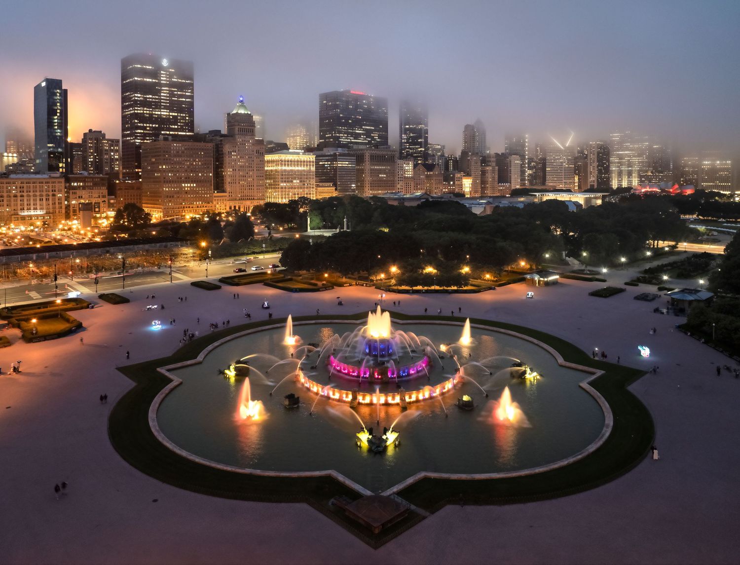A foggy night skyline drone image of Chicago with Buckingham Fountain illuminated in the foreground.