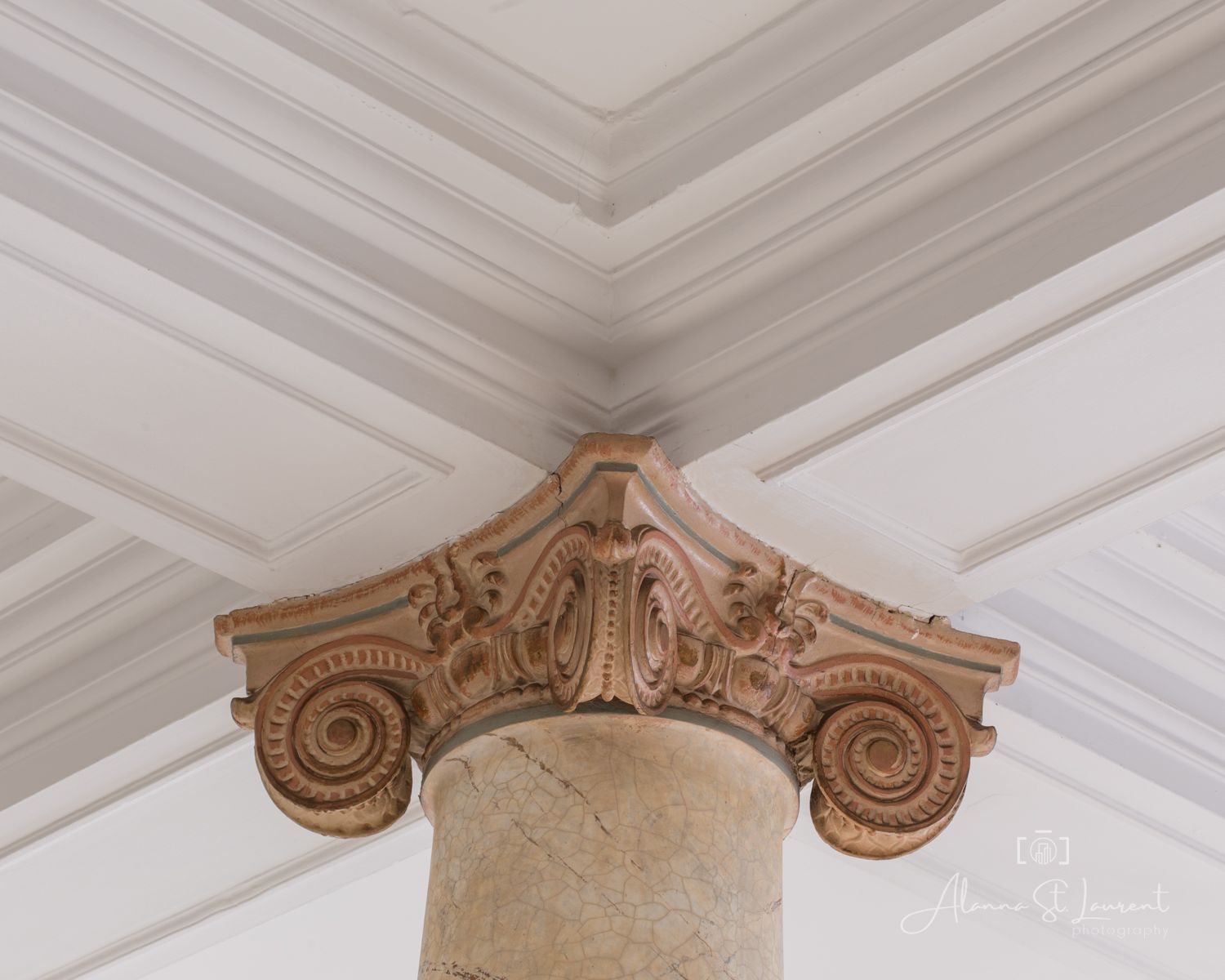Ornate Ionic column capital with scroll volutes and acanthus leaf details beneath a coffered ceiling.
