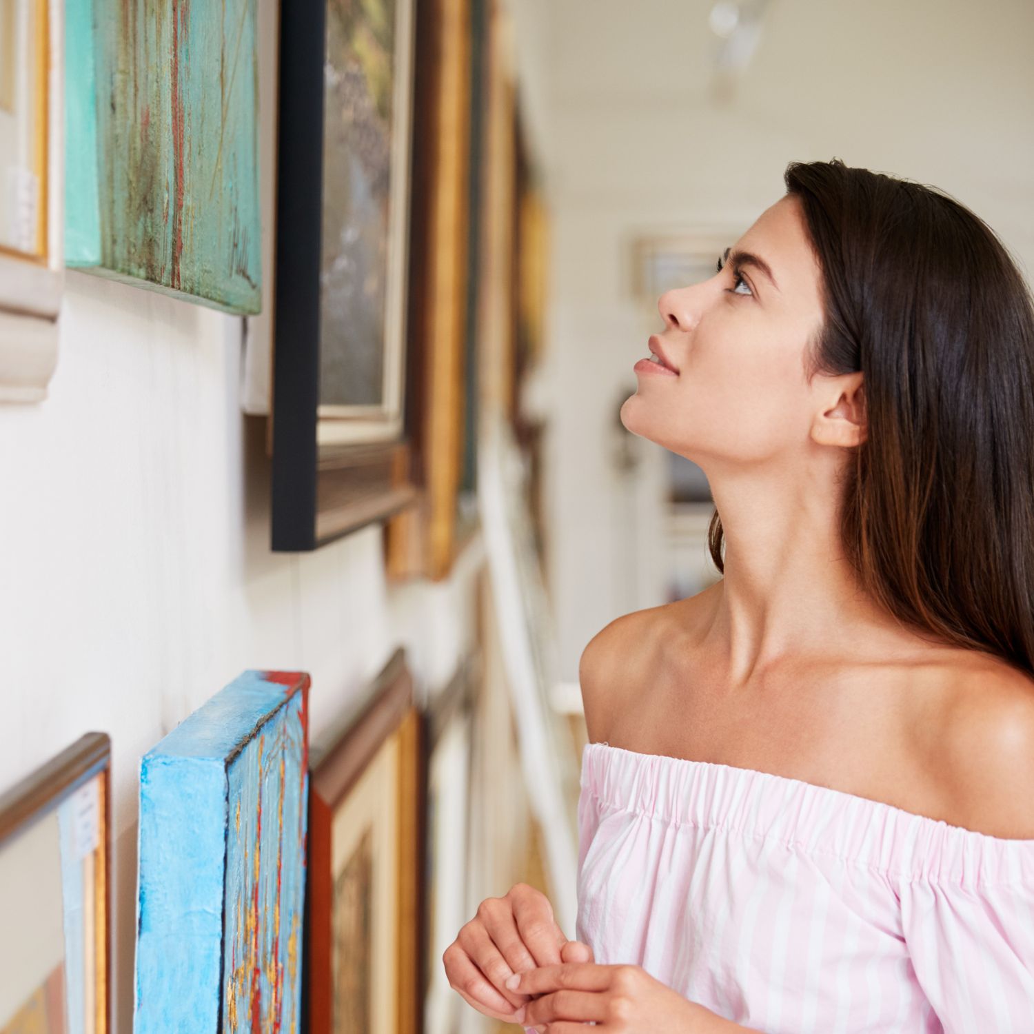 Young woman with dark hair admiring framed artwork on a gallery wall in a bright art studio.